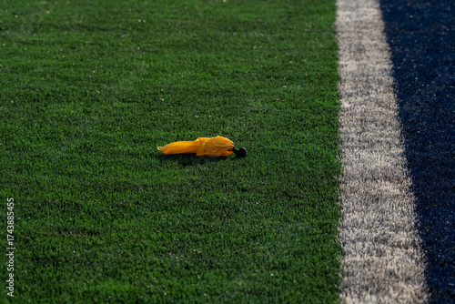 This minimalist photograph captures a bright yellow penalty flag lying on a vibrant green artificial turf sports field. 