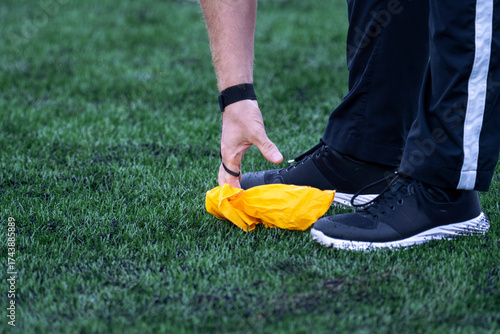 This close-up, low-angle photograph shows a game official's hand reaching down to grasp a bright yellow penalty flag resting on a vibrant green artificial turf field. 