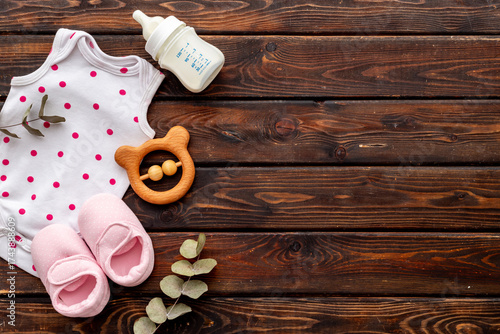 Flat lay of baby bodysuits and eco friendly accessories - booties, wooden rattle toy and baby bottle with milk on brown wooden background, top view