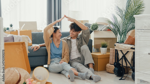 Fotografie Happy Asian couple sit on floor in front of sofa couch smile looking at camera making house sign in living room at new house