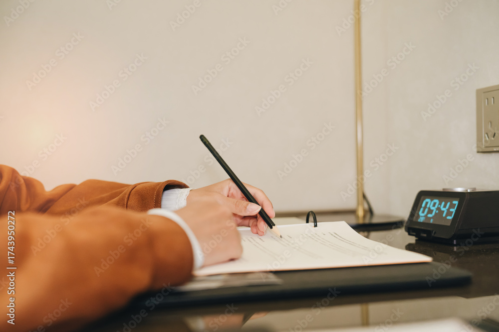 Fototapeta premium Person Writing Notes on Paper with Clock in Background in Modern Workspace Setting