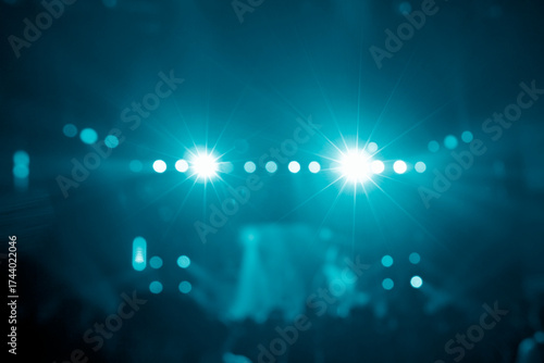 Bright teal stage lights with prominent starburst flares illuminate a dark, blurry background filled with numerous bokeh circles, suggesting a vibrant concert atmosphere or abstract light pattern.