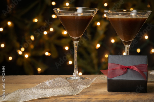 Two espresso martini cocktails and a present wrapped in black paper and red ribbon on a wooden table, Christmas tree in background.