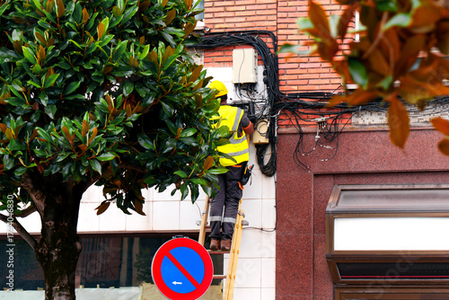 Electrician in yellow safety vest and helmet repairing electrical wiring on exterior wall of residential building using stepladder on city street