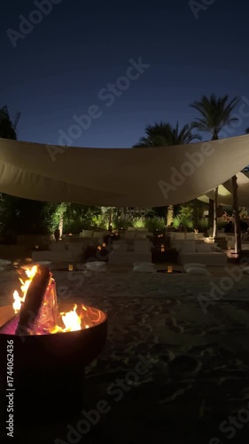 A night scene with a campfire, people sitting around, enjoying warmth and conversation. Tents and outdoor items visible, creating a cozy atmosphere. Dark sky, trees, and nature elements in the backgro