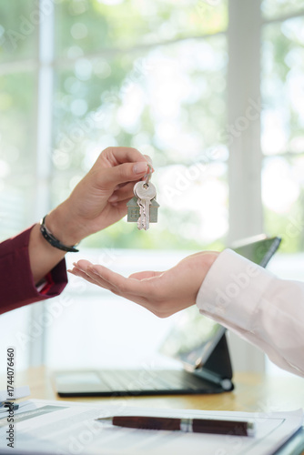 Smiling businesswoman holding calculator while discussing real estate financial planning with client, showing house model and documents on office desk.