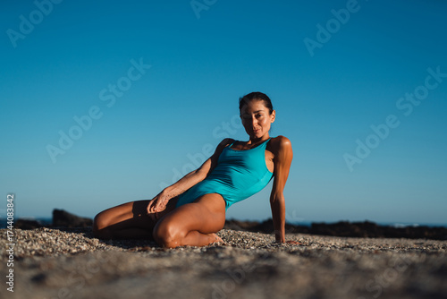 Smiling athletic woman in turquoise swimsuit relaxing on sandy beach, leaning back on her arms under morning sunlight, enjoying peaceful moment of rest after fitness workout near the ocean