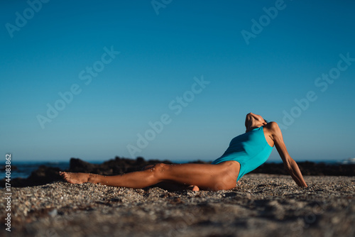 Fit woman in turquoise swimsuit relaxing on sandy beach at sunset, sitting gracefully with extended legs and arms behind her, enjoying peaceful seaside atmosphere under clear blue sky