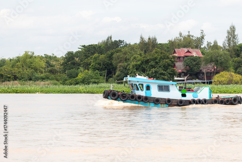 blue river tugboat cruising with black tire fenders near green riverbank and traditional house