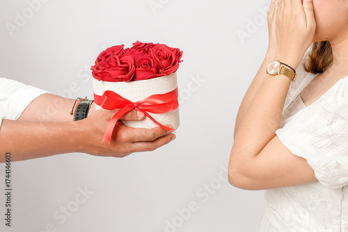 Close-up of a person presenting a round gift box filled with red roses and tied with a red ribbon to a surprised woman in white, symbolizing love, celebration, and romantic gestures.