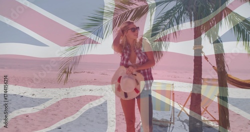 Standing in checkered shirt, shorts on sandy beach, with straw hat, palm tree, Union Jack flag