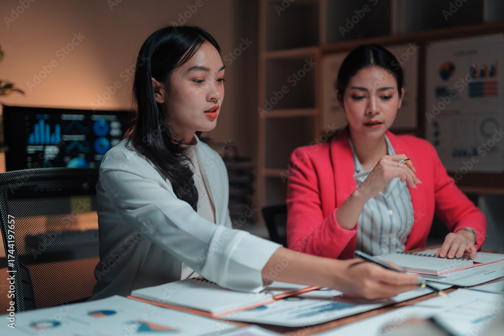 Fototapeta premium Two young Asian businesswomen working late in the office, discussing financial data and analyzing charts during a meeting