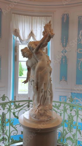 A majestic vertical full shot captures a white marble classical sculpture of two intertwined mythological figures positioned atop a pedestal on a grand interior staircase landing. The statue is