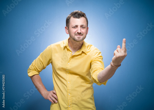 Smiling man giving the middle finger - showing fuck off gesture over blue background, dresses in yellow shirt. One guy with obscene gesture and looking to camera