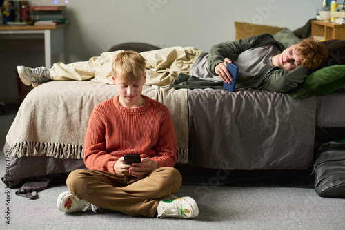 Caucasian teenage boy sitting on floor using smartphone while Caucasian teenage brother lying on bed holding phone, both engaging with devices in shared bedroom setting