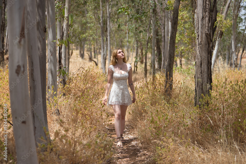 Naklejka premium Young woman, beautiful, blonde, wearing a floral dress, relaxed and calm, walking among the trees, in connection with nature and ecology, in an outdoor park. Concept of beauty, youth, millennial.