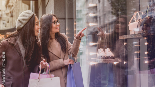 Two young women enjoy shopping while pointing finger in the shopwindow.