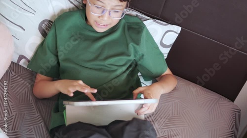 Asian girl focused on playing a game on a digital tablet at home. Top view shot shows childhood engagement with modern technology in her bed for enjoying her leisure time entertainment.