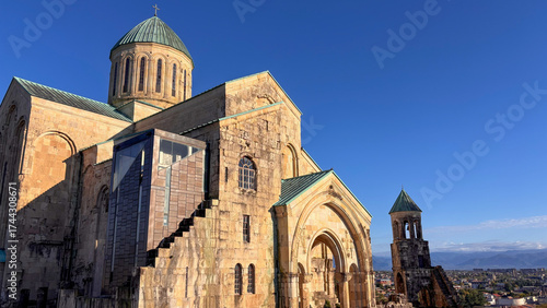 Bagrati Cathedral, 11th-century UNESCO World Heritage Site in Kutaisi, Georgia. Ancient stone architecture, dome & arches, blends with a modern glass & metal addition under a blue sky.