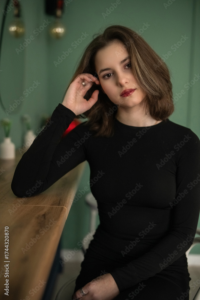 Fototapeta premium Young woman with shoulder-length hair, wearing a black top, gazes thoughtfully with soft lighting, capturing a moment of introspection and beauty in a serene atmosphere