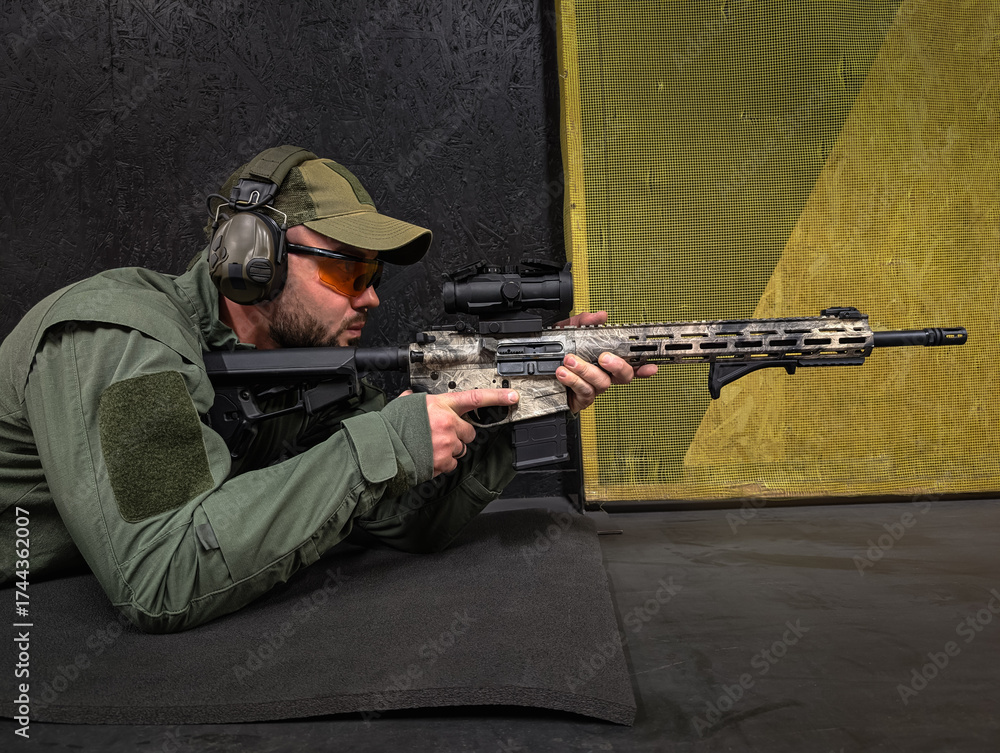Fototapeta premium A focused male shooter in tactical clothing, safety glasses, and earmuffs aims a modern camouflage rifle from a prone position.
