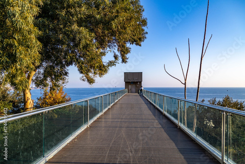 Fototapeta Naklejka Na Ścianę i Meble -  Modern pedestrian bridge with glass railings leading to Konyaaltı Beach, set against the Mediterranean Sea. A perfect blend of contemporary infrastructure and natural beauty in Antalya, Turkey.

