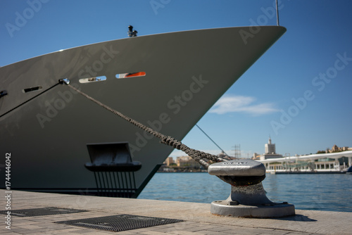 Large luxury yacht moored in marina, thick rope tied to metallic bollard on concrete dock under clear blue sky in bright sunlight, modern port background