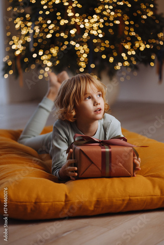 Wallpaper Mural Happy Caucasian girl, 9 years old, blond curly hair, in pajamas, lying on soft cushion holding gift box near Christmas tree with lights and white wall. Concept of holiday, New Year, childhood joy. Torontodigital.ca