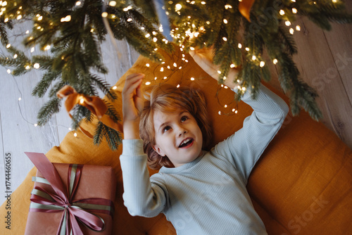 Happy Caucasian girl, 9 years old, blond hair, blue sweater, top view, lying on orange cushion under decorated Christmas tree with lights and gift. Concept of holiday, family, childhood, New Year.