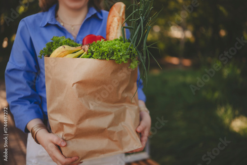woman carries brown paper bag with fresh vegetables and colorful fruits in lush garden, enjoying sunny afternoon.