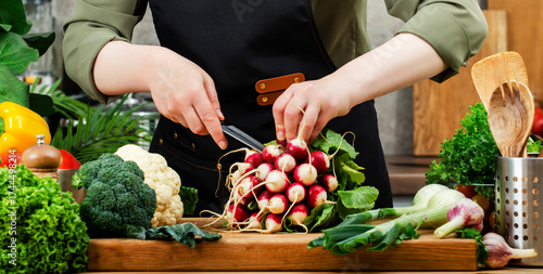 In bright kitchen, private chef chops fresh radishes and colorful vegetables, herbs onwooden cutting board