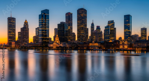 Modern city skyline at sunset reflecting in river with illuminated skyscraper and boat light trails. Urban development and cityscape concept.