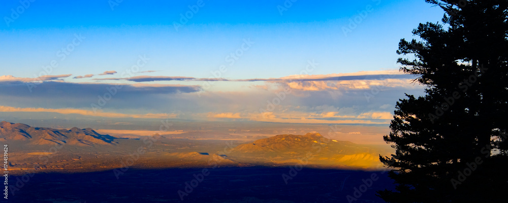Fototapeta premium A breathtaking sunset paints the sky with warm colors as the sun sets behind the mountains. Looking west from Sandia Peak Albuquerque New Mexico. Trees stand tall, silhouetted against the vibrant hor