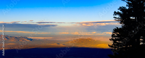 A breathtaking sunset paints the sky with warm colors as the sun sets behind the mountains. Looking west from Sandia Peak Albuquerque New Mexico.  Trees stand tall, silhouetted against the vibrant hor