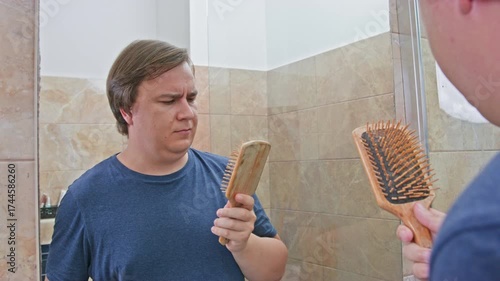 A man in a blue shirt closely examines a hairbrush while looking at his reflection in a bathroom mirror, pondering grooming habits. noticing the first hair loss when going bald