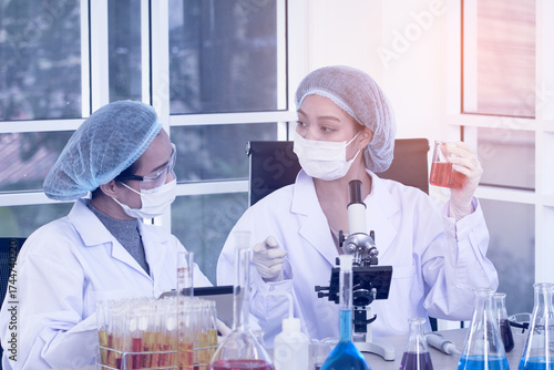 Two female scientists in lab coats and protective gear are performing an experiment, holding a flask with red liquid while observing and taking notes on a tablet.