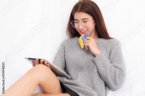 Happy Asian woman in glasses holding a tablet and credit card enjoying online shopping and e-commerce from white room.