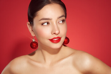 Portrait of beautiful young woman on red background, red lipstick and festive makeup, Christmas toy earrings, shiny glowing skin, Winter holidays concept