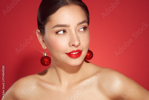Portrait of beautiful young woman on red background, red lipstick and festive makeup, Christmas toy earrings, shiny glowing skin, Winter holidays concept