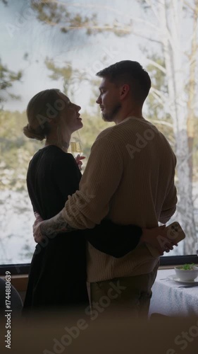 An elegant young loving couple enjoying romantic date with champagne. A man and a woman standing by window on a sunny day, looking beautiful winter landscape. Celebrating moment. Vertical video.