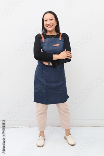 Full body shot of smiling Asian waitress in blue apron, looking at camera, standing confidently, arms crossed on chest against white background.
