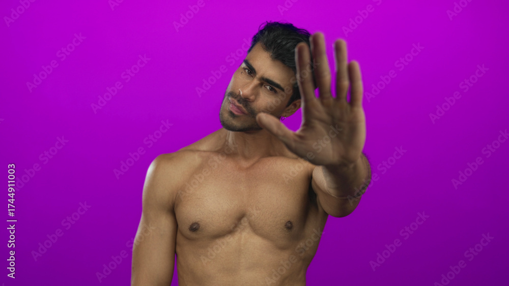 Fototapeta premium Young hispanic man with firm expression pushing palm forward across bare chest in purple studio; stop refusal defiance.