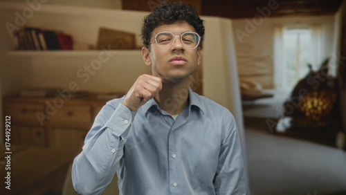 Canvas Print Man wearing glasses holds fist to chest in building interior with warm light and blurred furnishings; determination