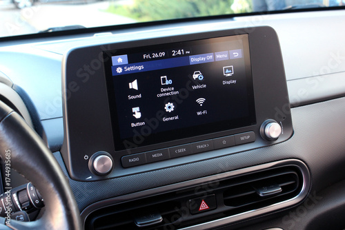 Car infotainment touchscreen displaying settings menu with icons for sound, Wi-Fi, and vehicle options. Dashboard features matte black finish with silver knobs and control buttons. Car multimedia.