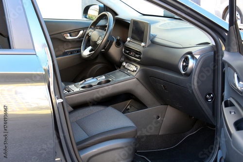 Electric car driver seat. Electric car modern interior features black leather steering wheel and dark fabric seats. An open drivers door reveals an elegant dashboard and ergonomic center console. 