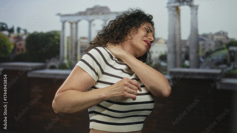 Fototapeta premium Woman in colorful striped top grasps neck and rubs shoulder beside historic stone building at roman ruins; relief.