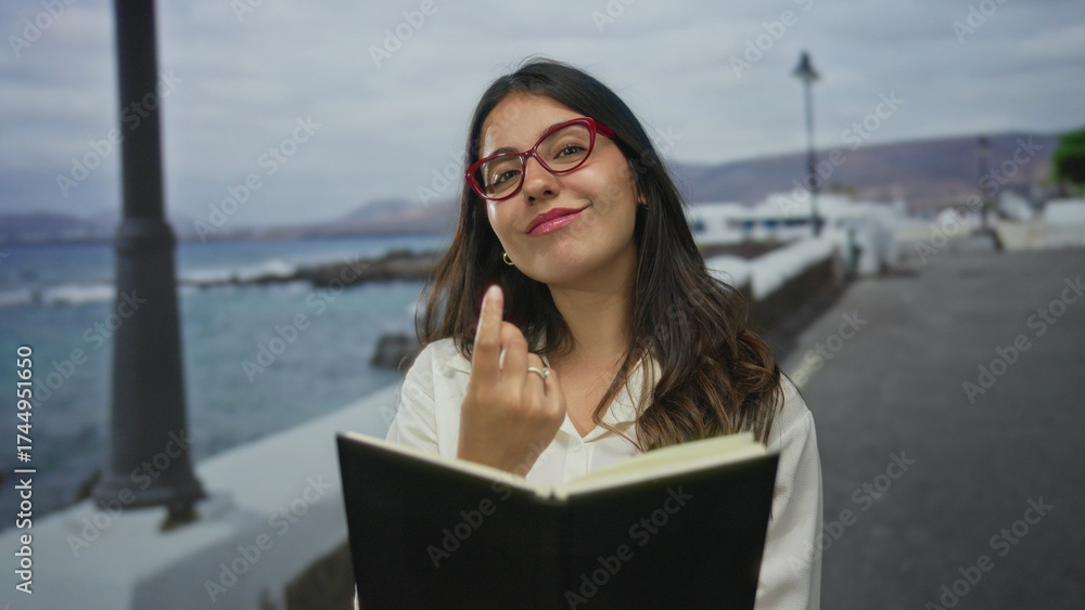 Fototapeta premium Young hispanic woman with red glasses reading black notebook on seaside promenade by gentle ocean waves; tranquility.