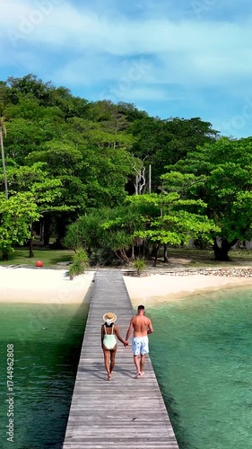 A couple strolls along a scenic wooden pier towards a lush landscape at Koh Kham Island, Thailand. a couple of caucasian men and Asian women on vacation