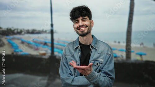 Young man smiling at beach with sea background under a cloudy sky, wearing denim jacket, capturing outdoor leisure and happiness in a seaside atmosphere.