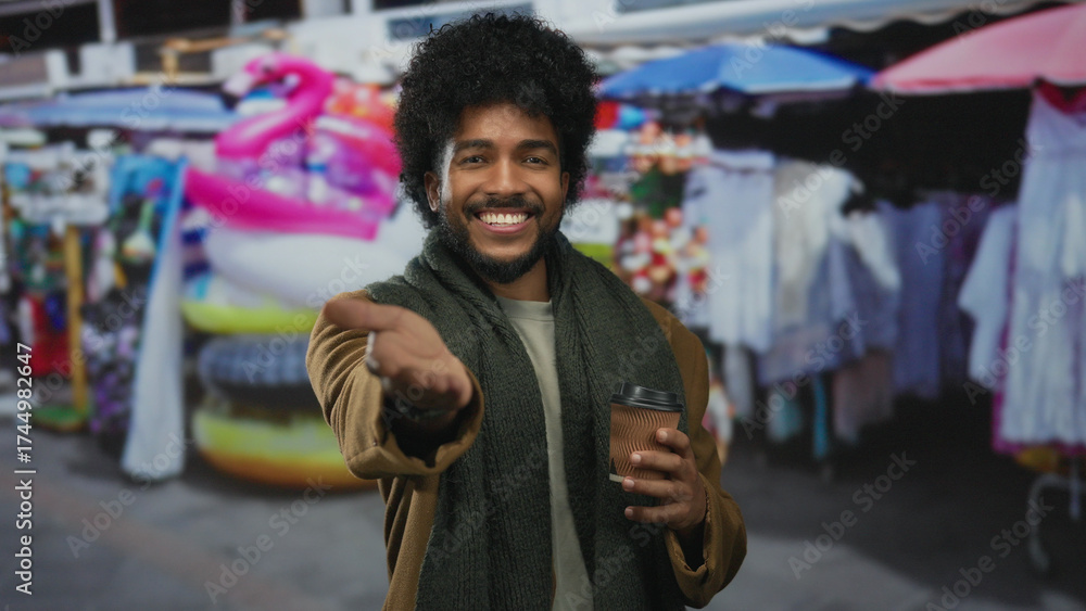 Fototapeta premium Smiling man in market street holding coffee with vibrant stalls in background reaching playfully forward.